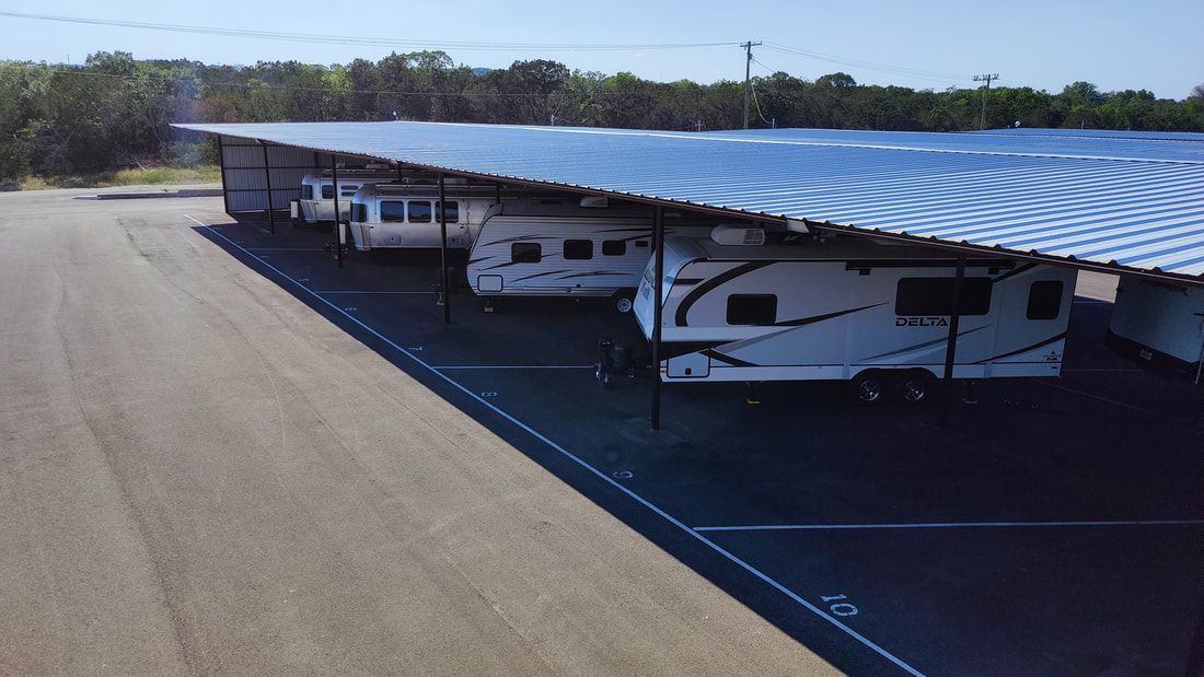 RV storage units with trailers parked under a metal roof, asphalt lot, trees in background.