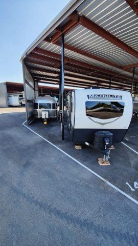 RV parked under a metal carport beside an industrial building, with a trailer hitch in front.
