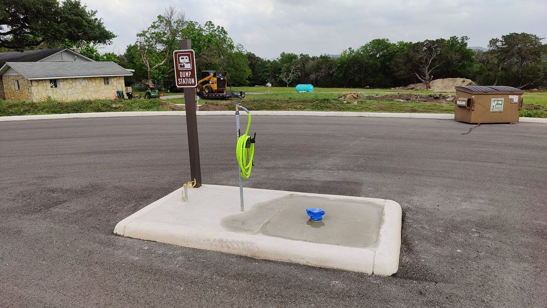 Campsite water station: concrete base with faucet, hose, and sign. Asphalt surface, trees, and buildings in the background.