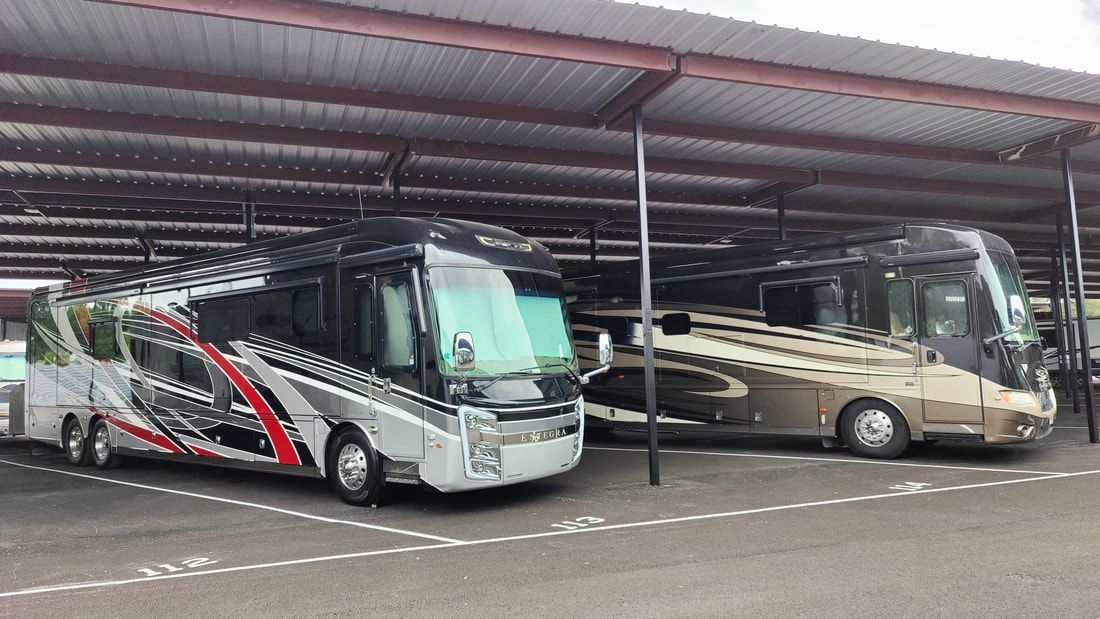 Two RVs parked under a brown metal awning. The RVs are silver with colorful graphics and tan.