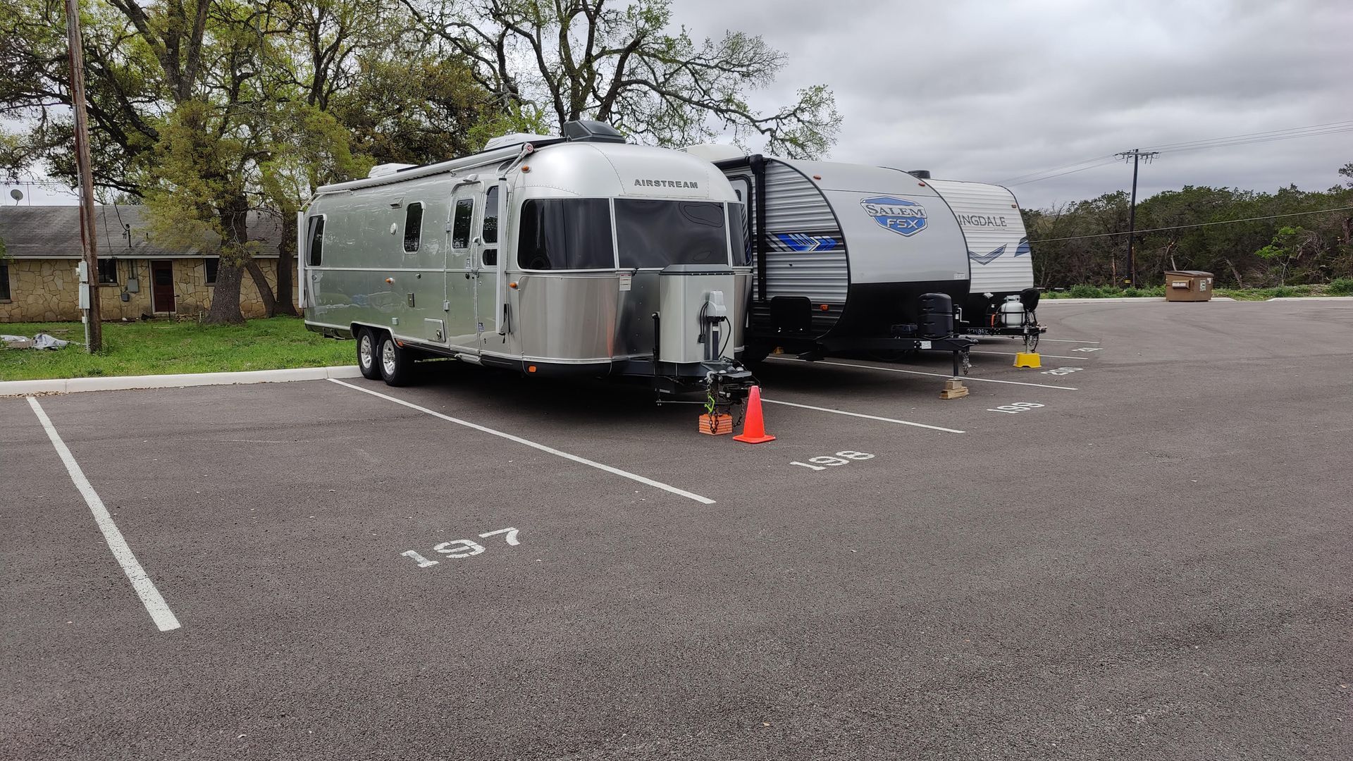 Silver RV and blue travel trailer parked in a lot, with orange cones between them on an overcast day.