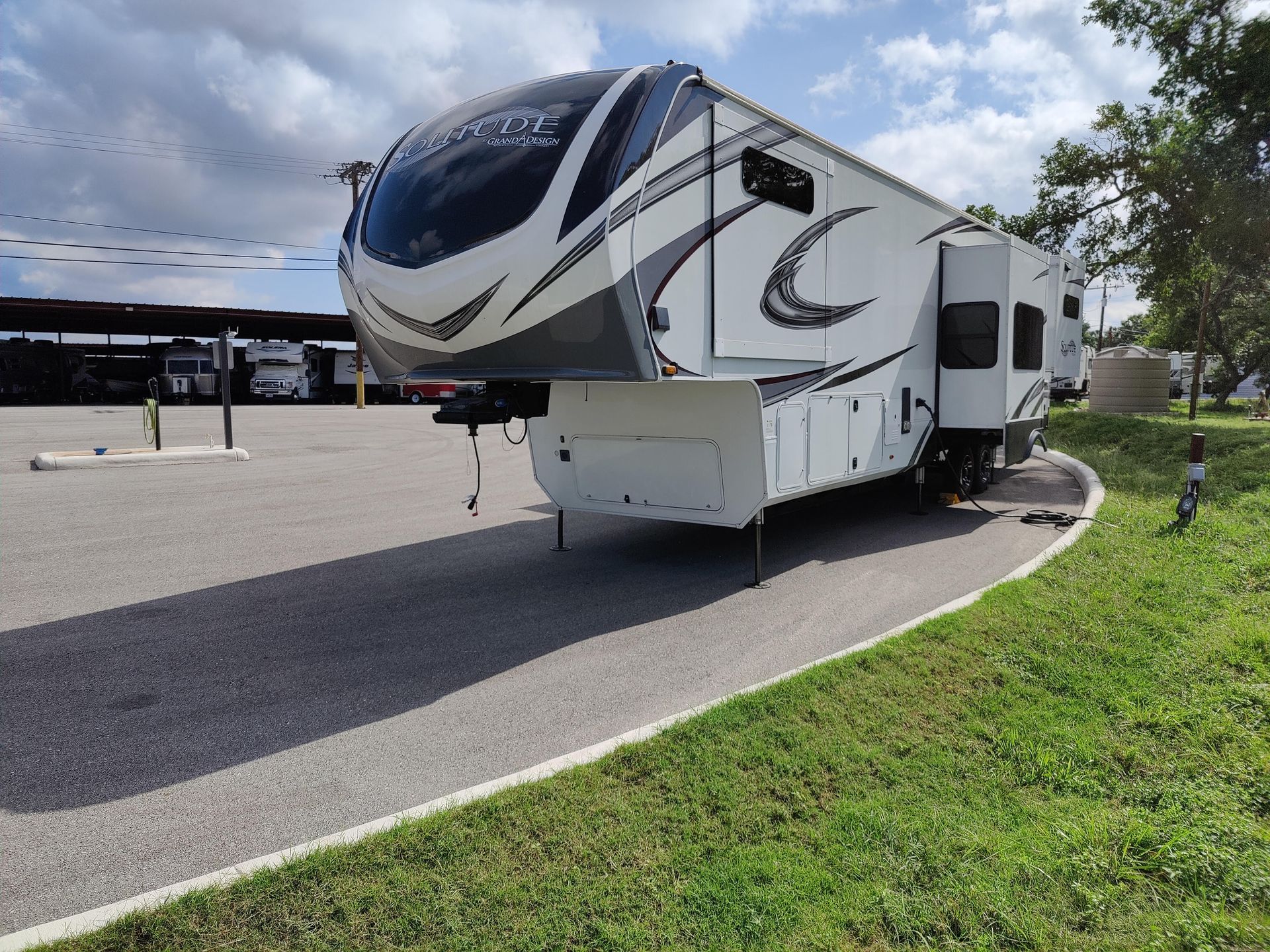 White fifth-wheel RV parked on a paved lot beside a grassy curb under a cloudy sky