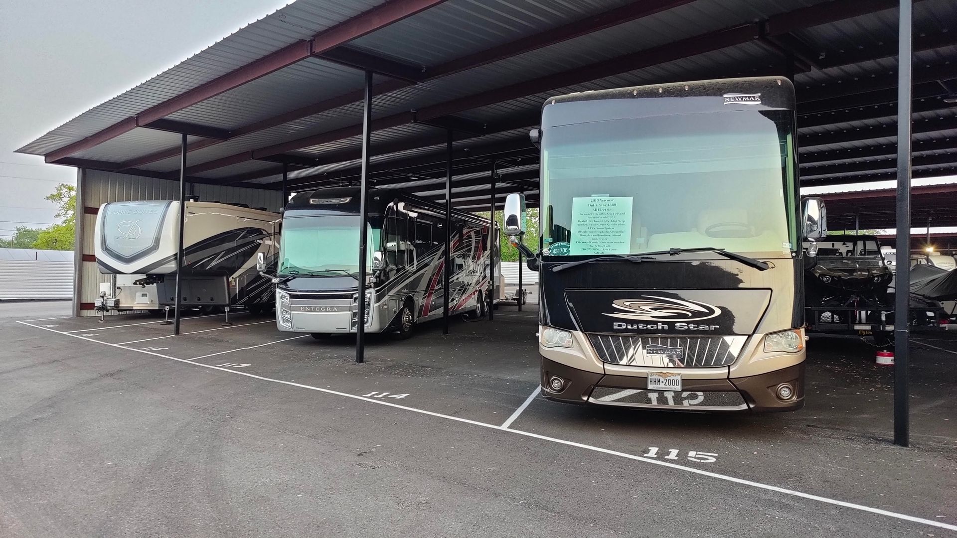 Covered parking lot with several motorhomes and RVs parked in a row, including a large tan coach in front.