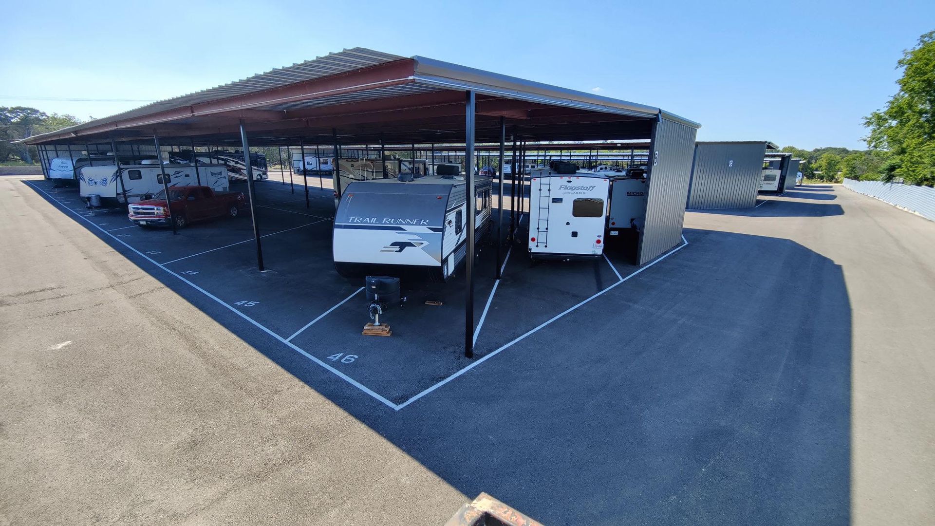 Covered parking lot with two white utility trailers under a metal canopy, marked parking spaces around them