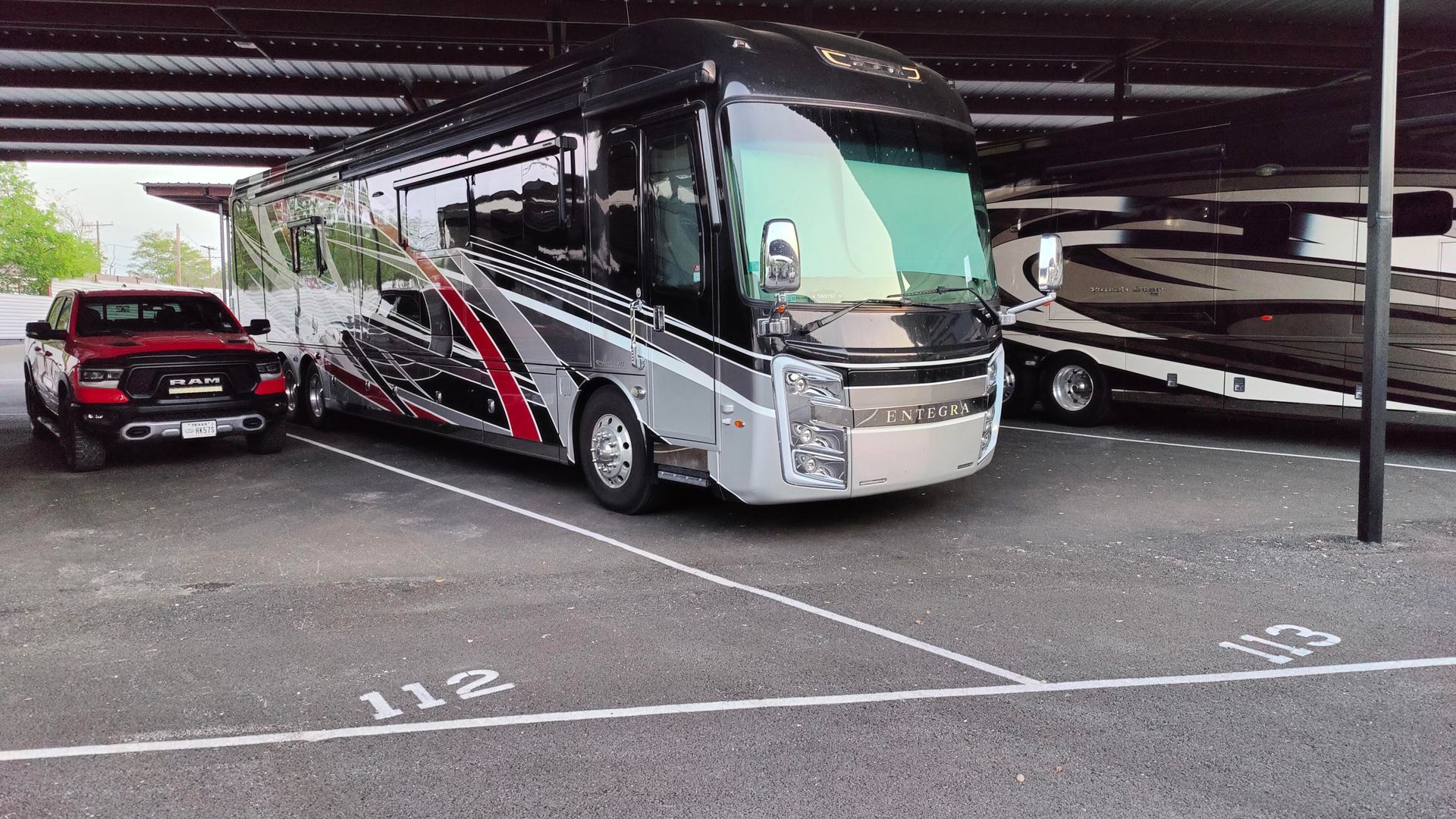 Silver and black motorcoach parked in a covered lot beside other RVs and marked parking spaces