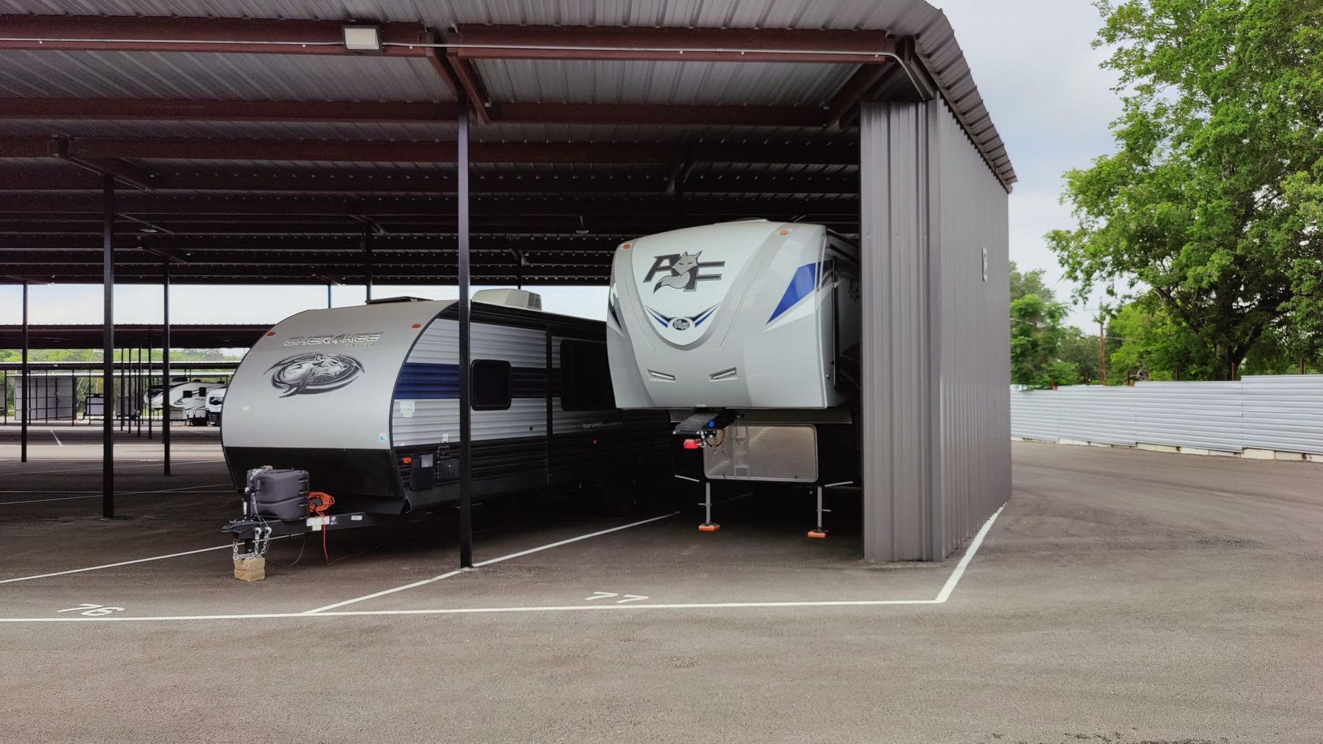 Two travel trailers parked under a metal carport, with an open paved lot and trees nearby.