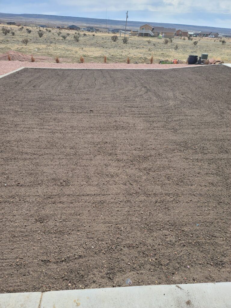 Gravel-covered lot bordered by dirt, with a desert landscape and distant buildings under a clear sky.