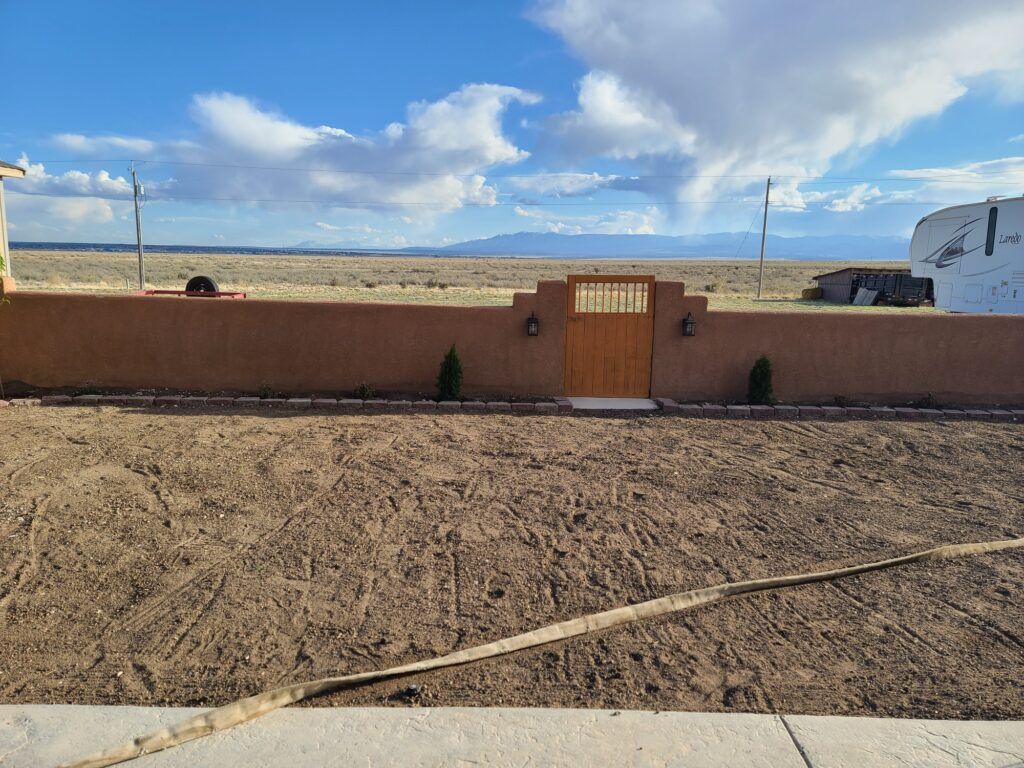 Adobe wall and gate facing a sandy yard with mountains and a blue sky in the background
