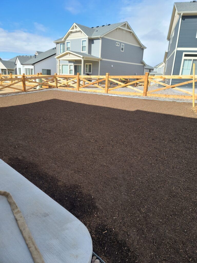 Newly spread dark mulch in a yard, with a sidewalk and suburban houses under a blue sky.