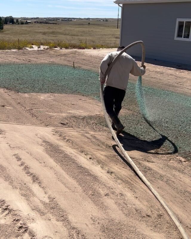 Worker spreading gravel beside a house on a dirt lot with a hose on the ground