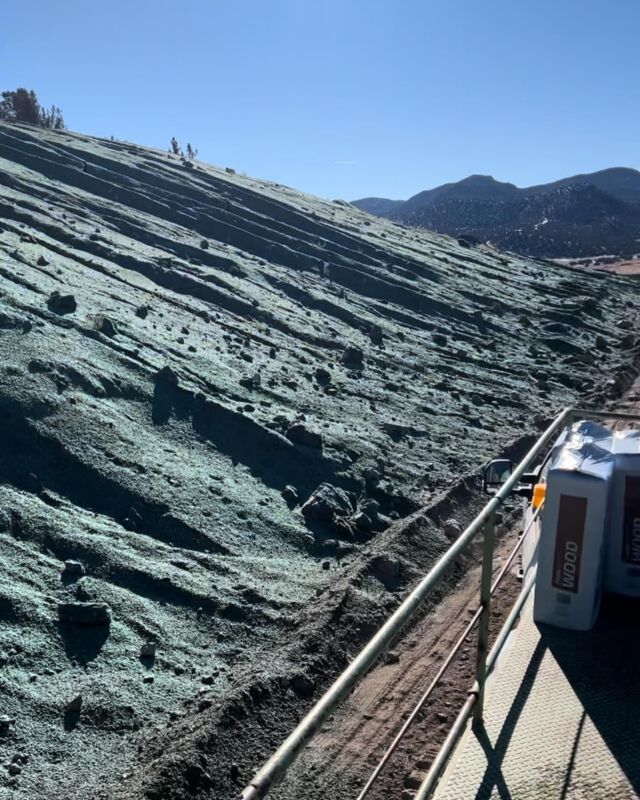 Steep greenish hillside beside railway tracks, with mountains in the distance under a blue sky.