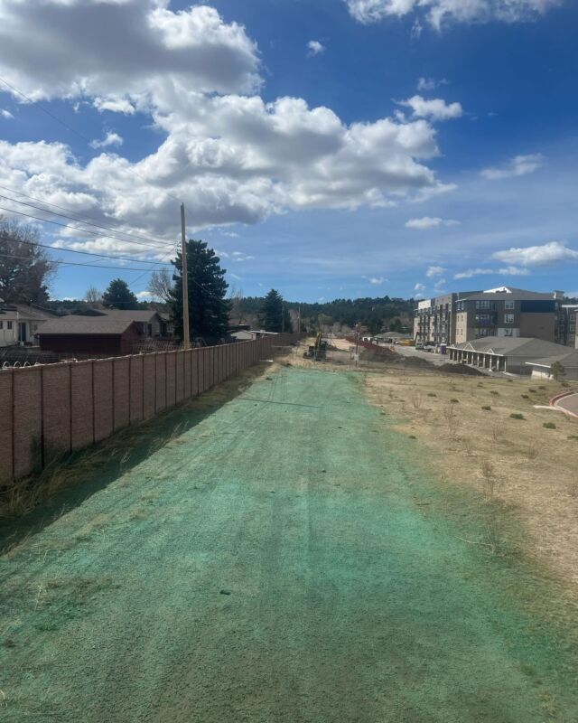 Green-covered road under construction between houses and apartments beneath a cloudy blue sky