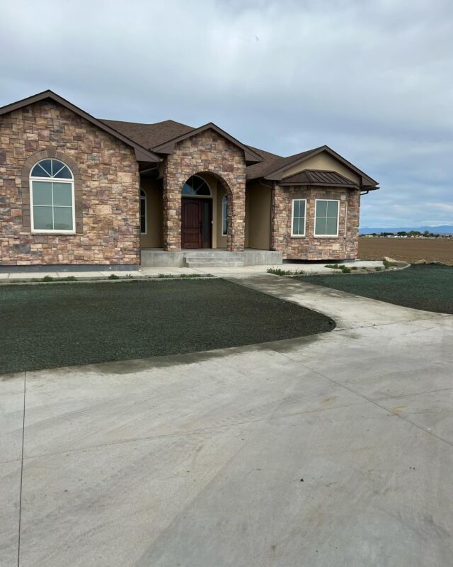 Single-story brick house with arched entry, dark roof, and a wide concrete driveway under a cloudy sky
