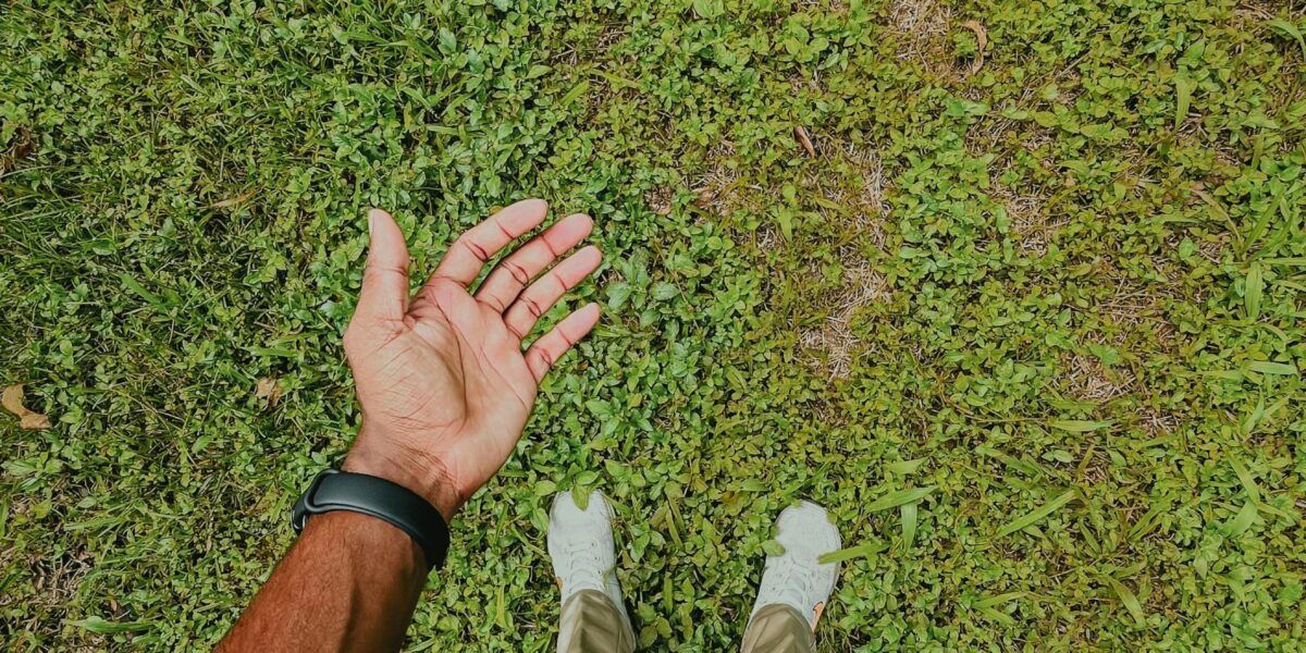 Hand resting on green grass beside white sneakers and a black wristband