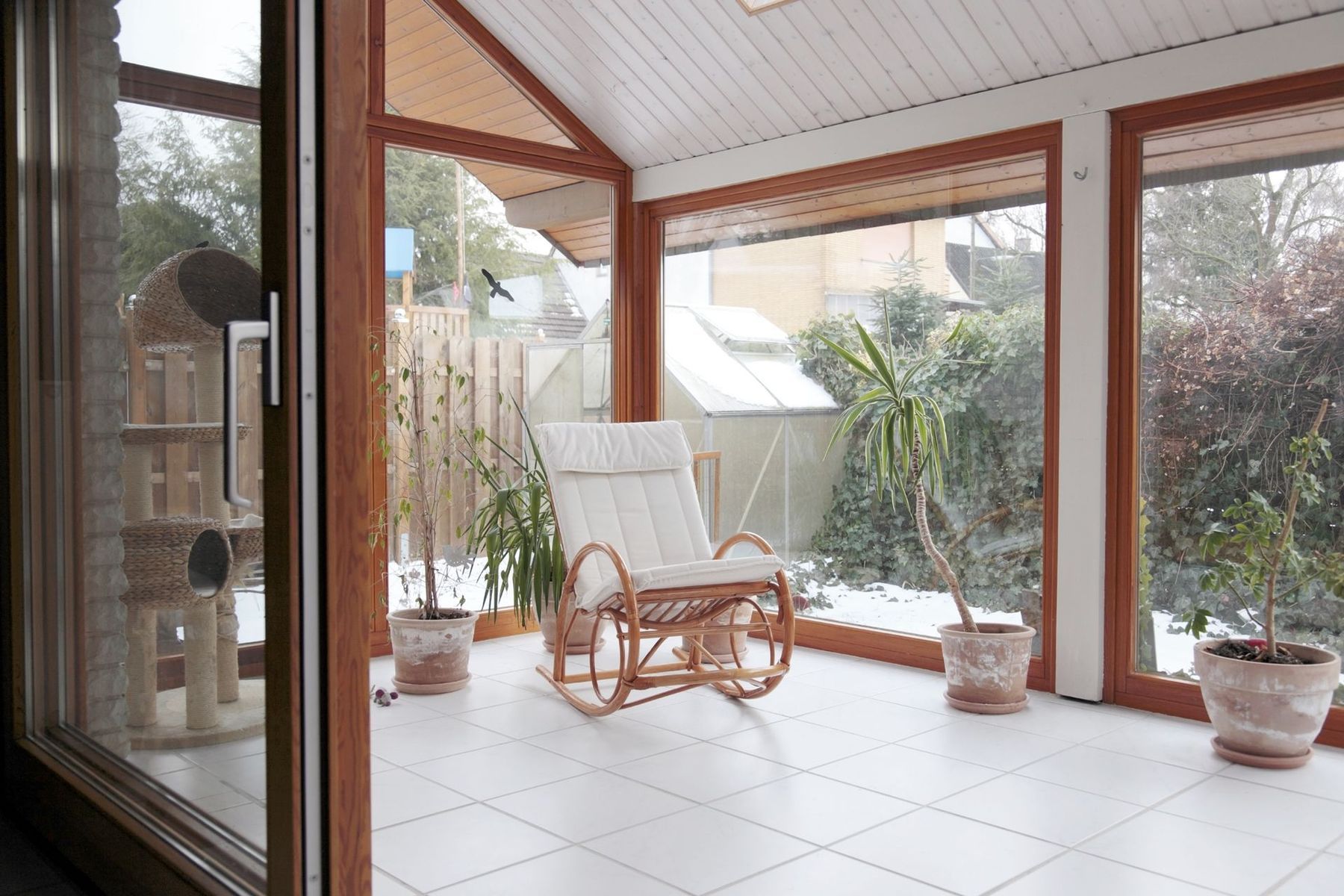 A sunroom with white tiled floors, a rattan rocking chair with a white cushion, several potted plants, and snow outside.