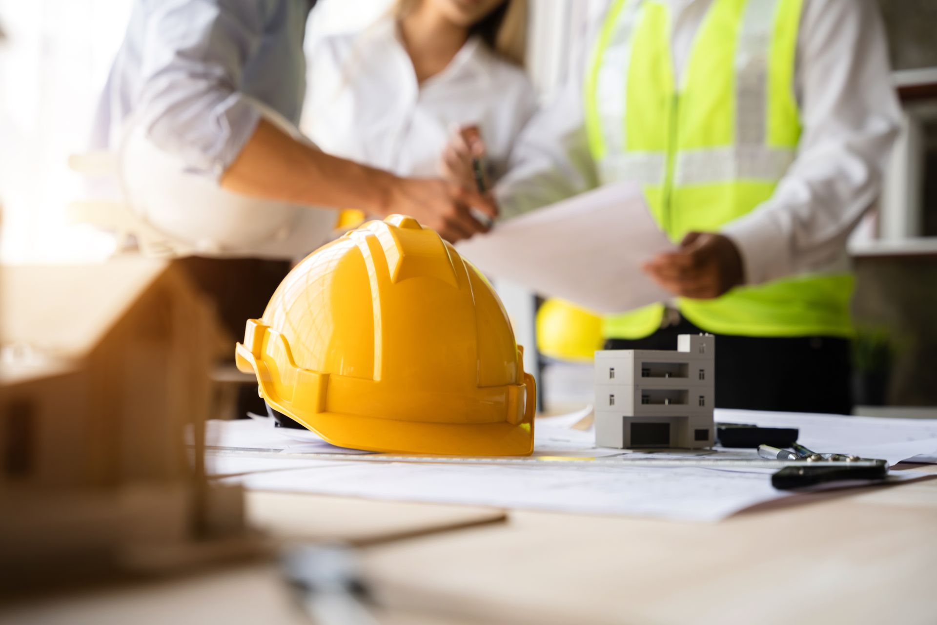 A yellow hard hat and small model building rest on construction blueprints as three people collaborate in the background.