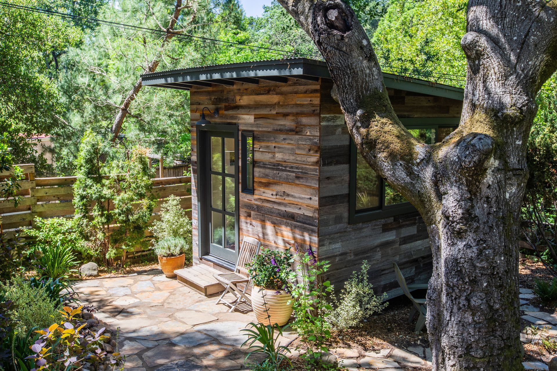A small, rustic wooden cabin with a dark door and window sits on a stone patio in a sunny, tree-filled garden.