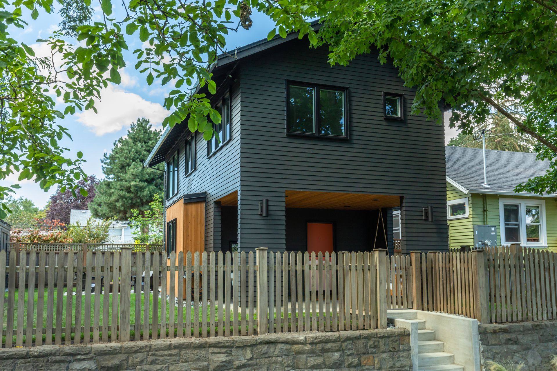 A modern, two-story house with black horizontal siding, a natural wood accent, and a front porch, behind a wooden fence.