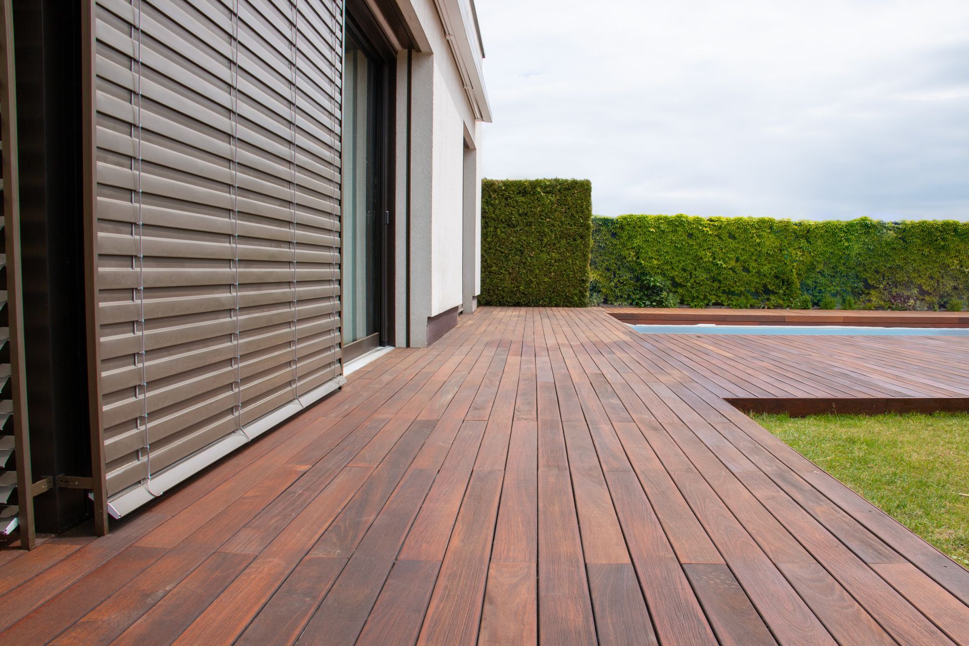 A brown wood deck extends from a modern house toward a small backyard pool and a neatly trimmed hedge.