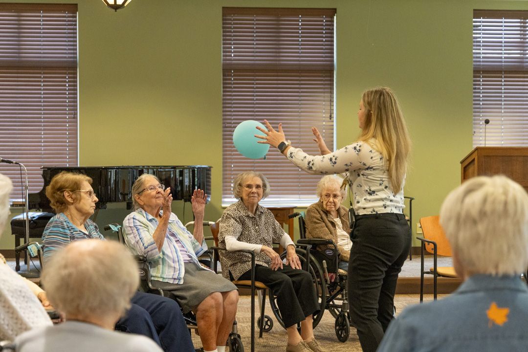 A woman is holding a balloon in front of a group of elderly people in chairs.