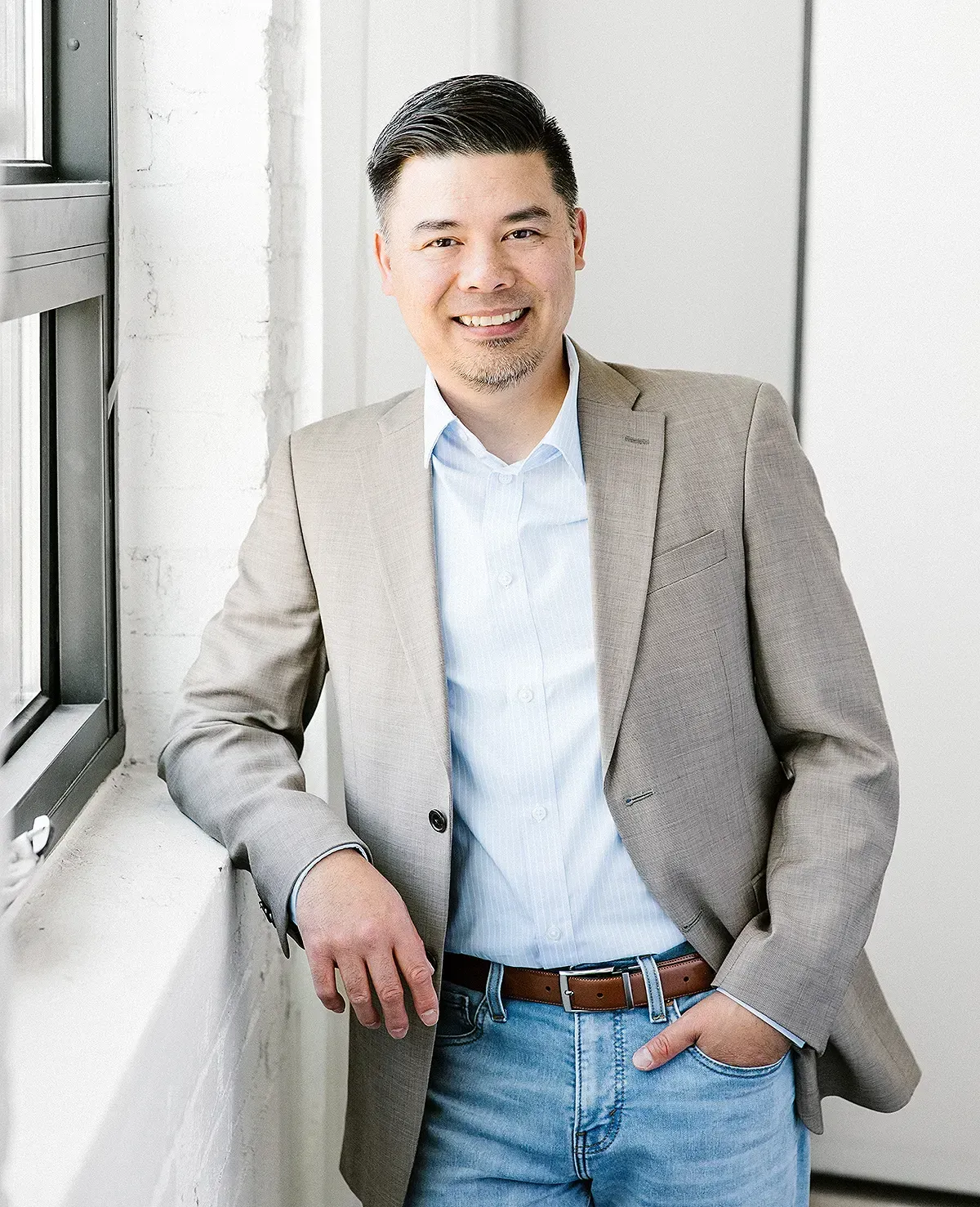 A man, Danny Truong, in a suit and jeans is leaning against a window.