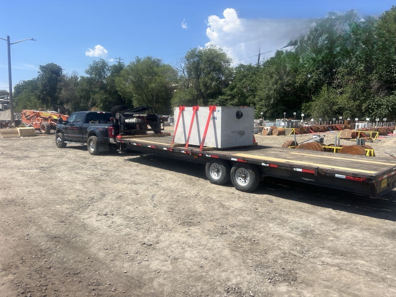 Flatbed trailer with a white crate parked on a dirt lot under a blue sky, with trees and equipment in the background