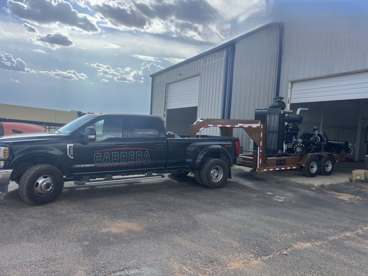 Black pickup truck hitched to a trailer in front of an industrial warehouse under a cloudy sky