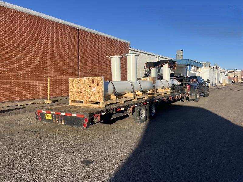 Flatbed trailer loaded with pipes and equipment parked beside a brick building in an industrial lot