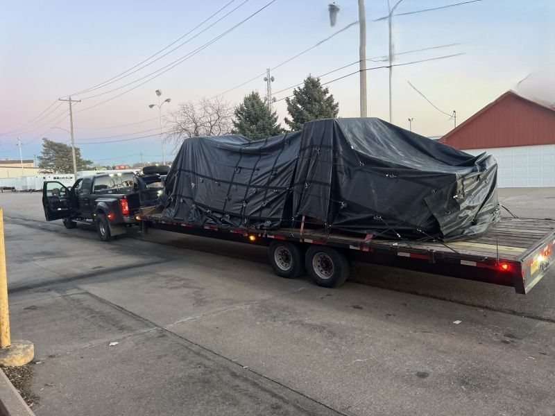 Black pickup towing a flatbed trailer loaded with a tarp-covered vehicle on a street at dusk