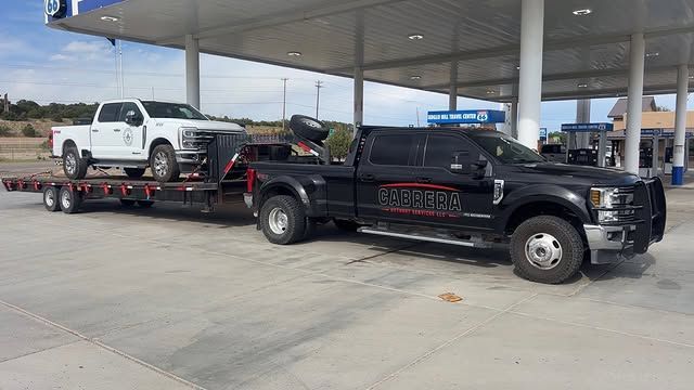 Black tow truck hauling a white pickup on a flatbed at a gas station