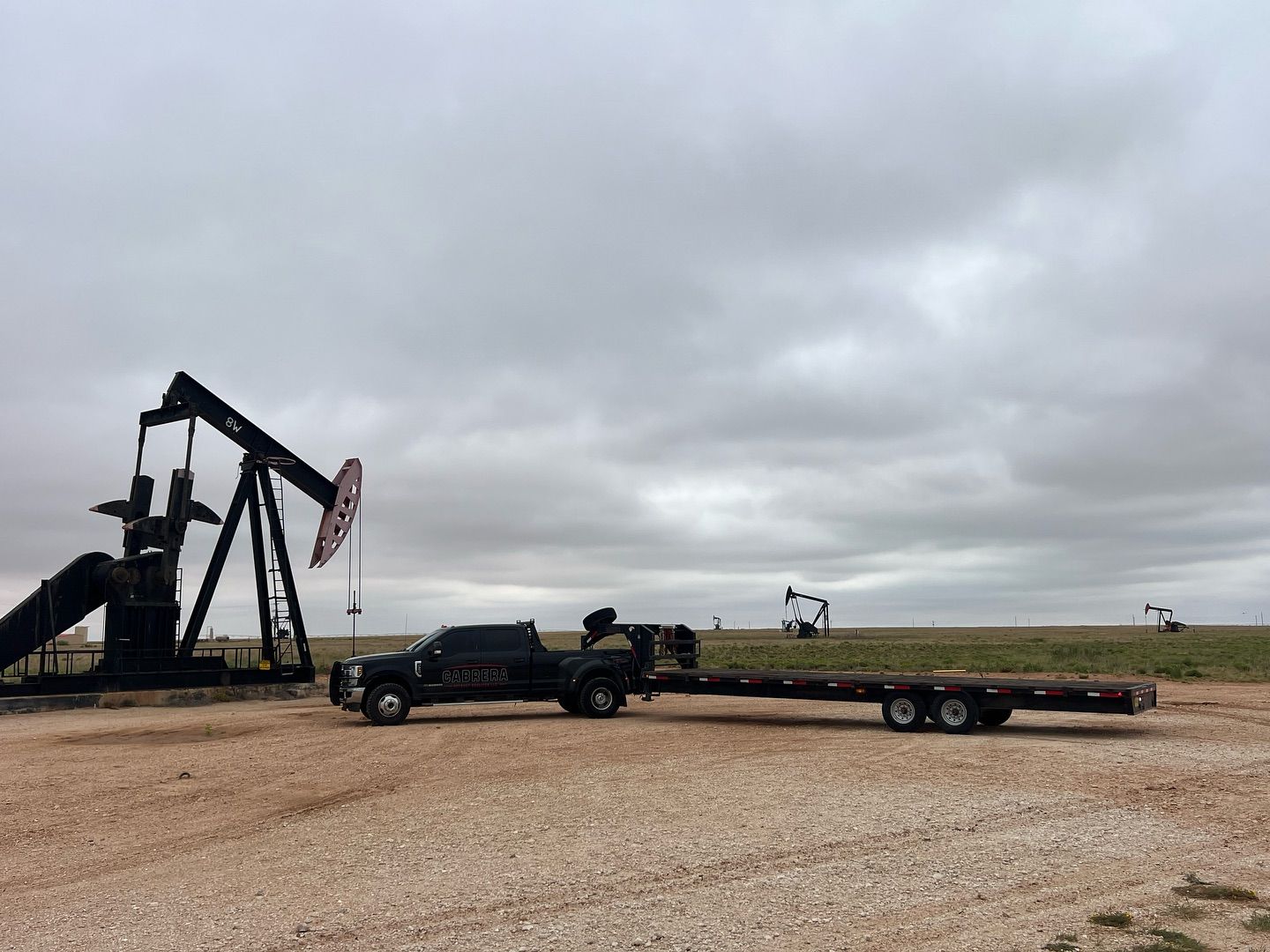 Black pickup hauling a long trailer beside a large oil pumpjack under a cloudy sky