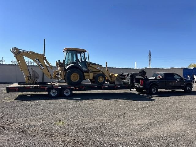 Black pickup towing a flatbed trailer carrying a backhoe on a gravel lot under a clear blue sky