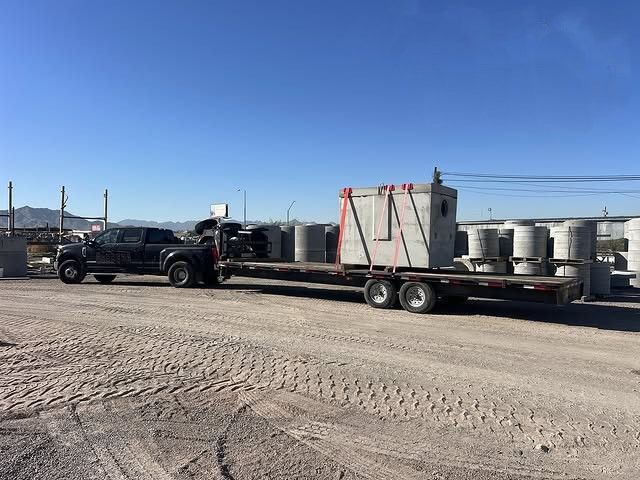 Black pickup towing a flatbed trailer loaded with large concrete pipes in a gravel lot