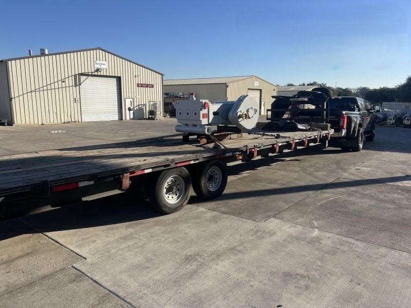 Flatbed trailer with heavy equipment parked on a concrete lot beside beige industrial buildings.