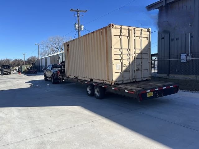 Flatbed trailer carrying a large beige shipping container in an industrial lot