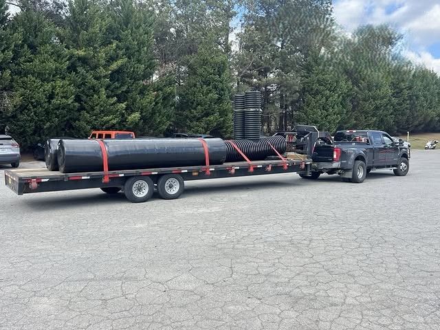 Truck towing a flatbed trailer loaded with a large black pipe coil in a parking lot
