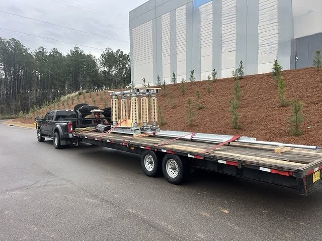 Black pickup towing a flatbed trailer loaded with long metal beams on a wet roadside near a building.