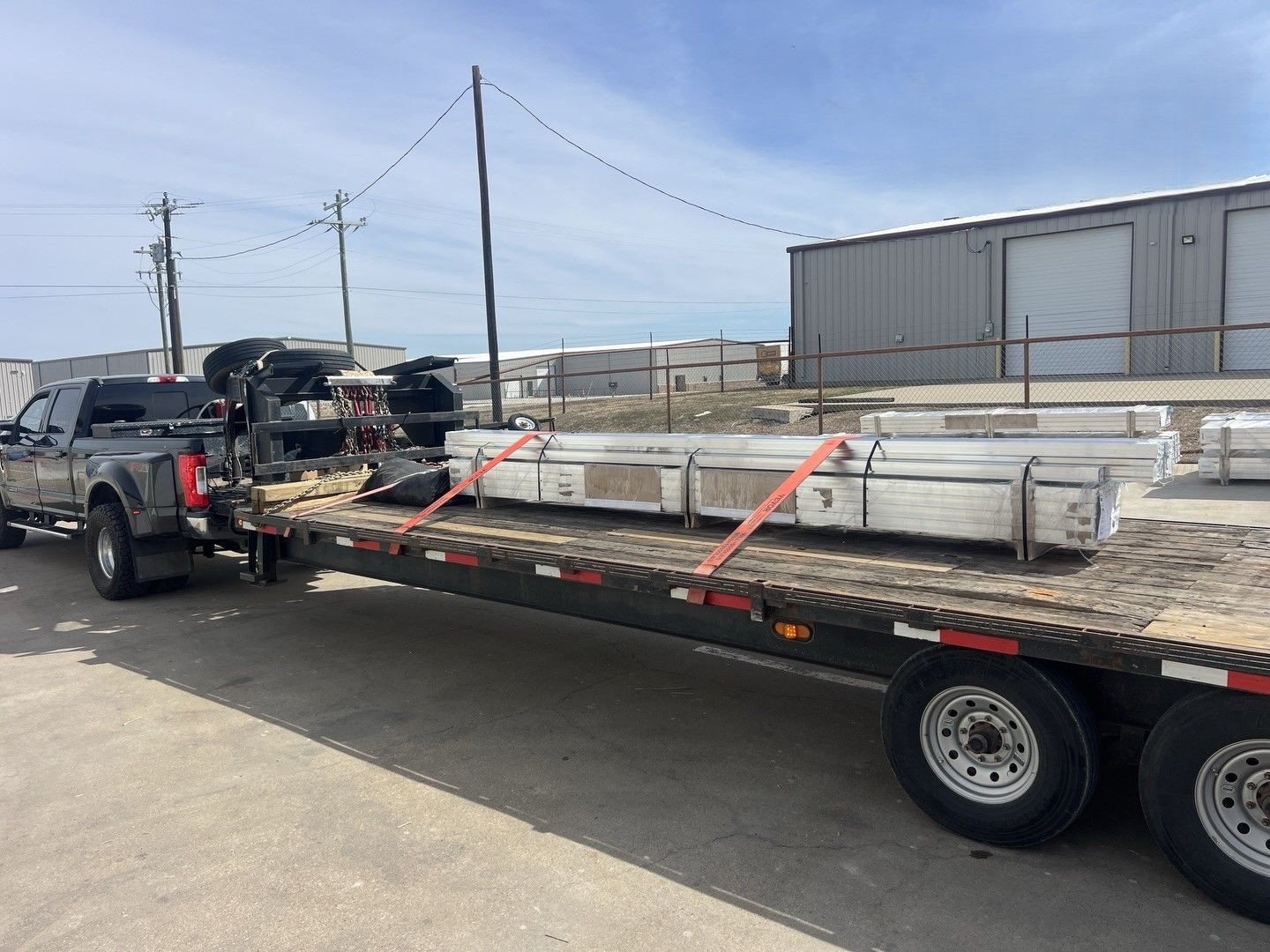 Flatbed trailer carrying long white beams, parked in an industrial lot beside a building