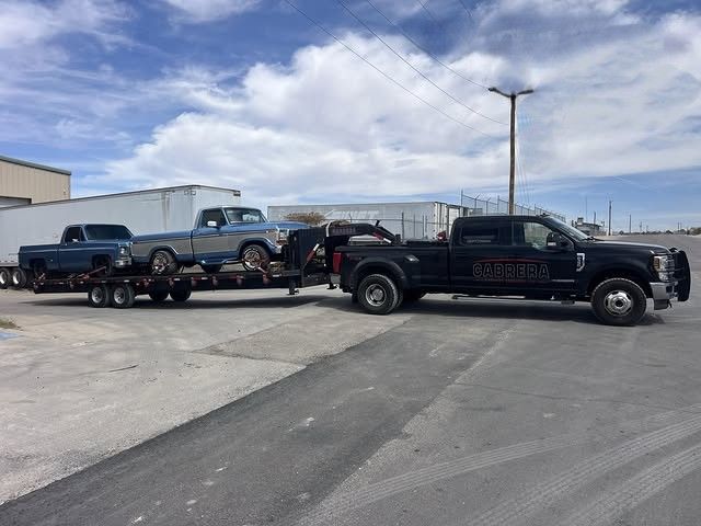 Black pickup truck towing a flatbed trailer with vehicles across a paved lot under a cloudy sky