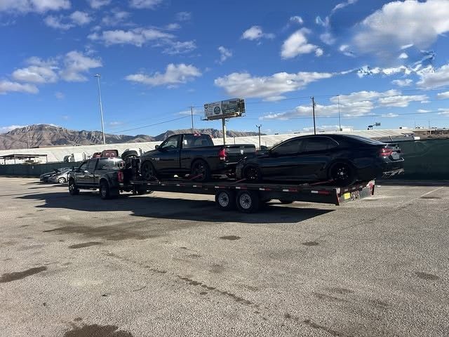 Black car on a trailer in a parking lot under a bright blue sky, with a pickup truck hitched in front.