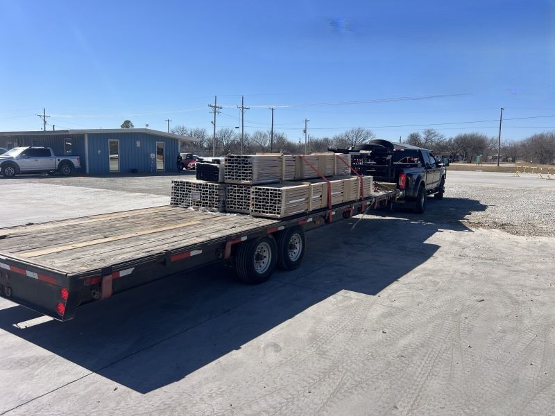 Flatbed trailer loaded with stacked pallets, parked in a sunny gravel lot beside a small building.