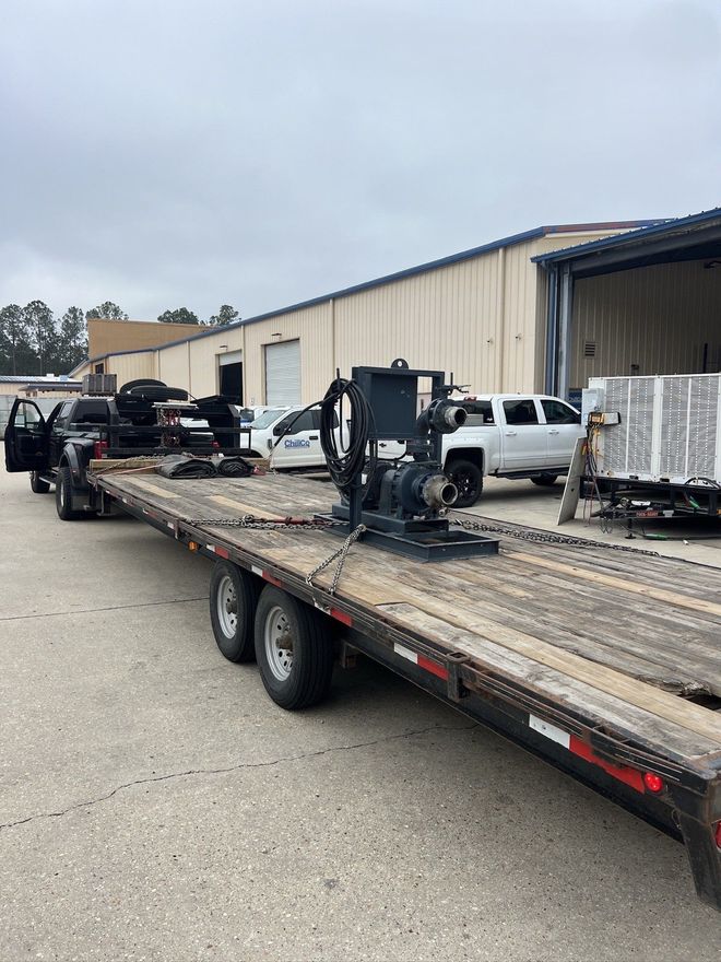 Flatbed trailer loaded with heavy equipment in an industrial parking lot beside a warehouse.