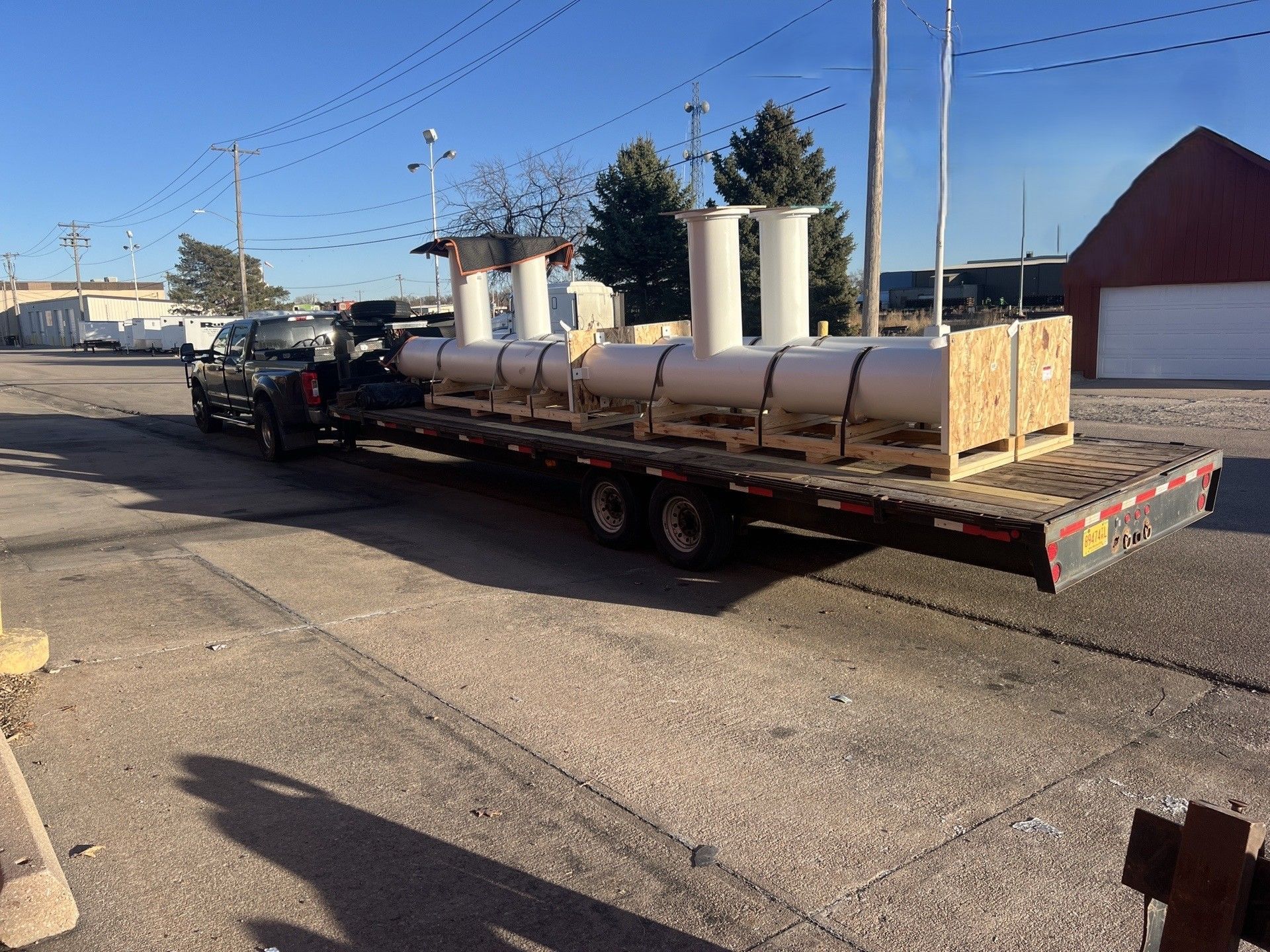 Flatbed trailer hauling large white tanks on a street beside industrial buildings under a clear blue sky