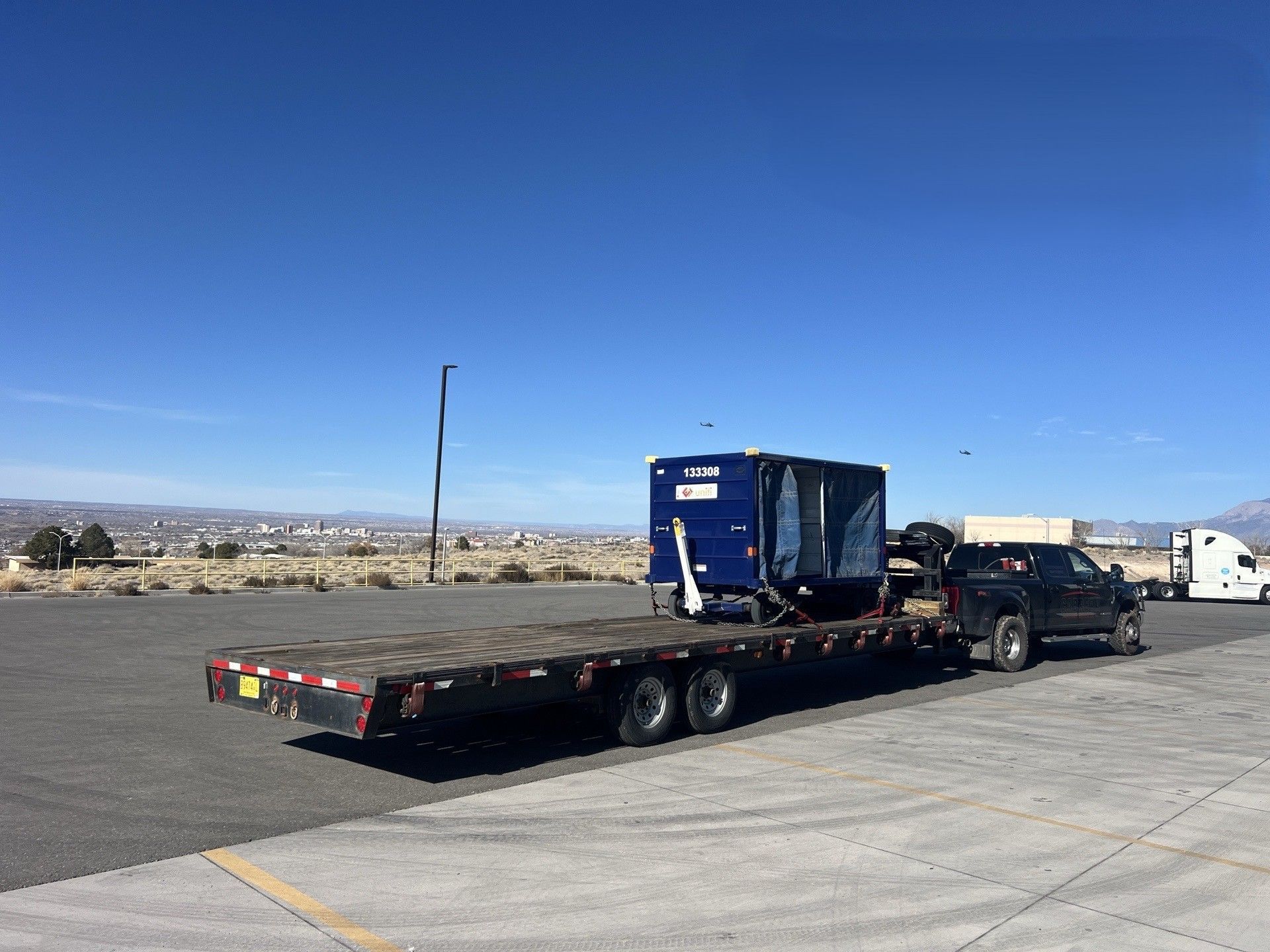 Flatbed tow truck carrying a blue machine in a parking lot under a clear sky