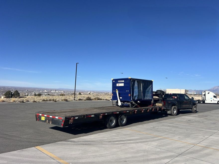 Flatbed tow truck carrying a blue machine parked on a wide roadside lot under a clear blue sky