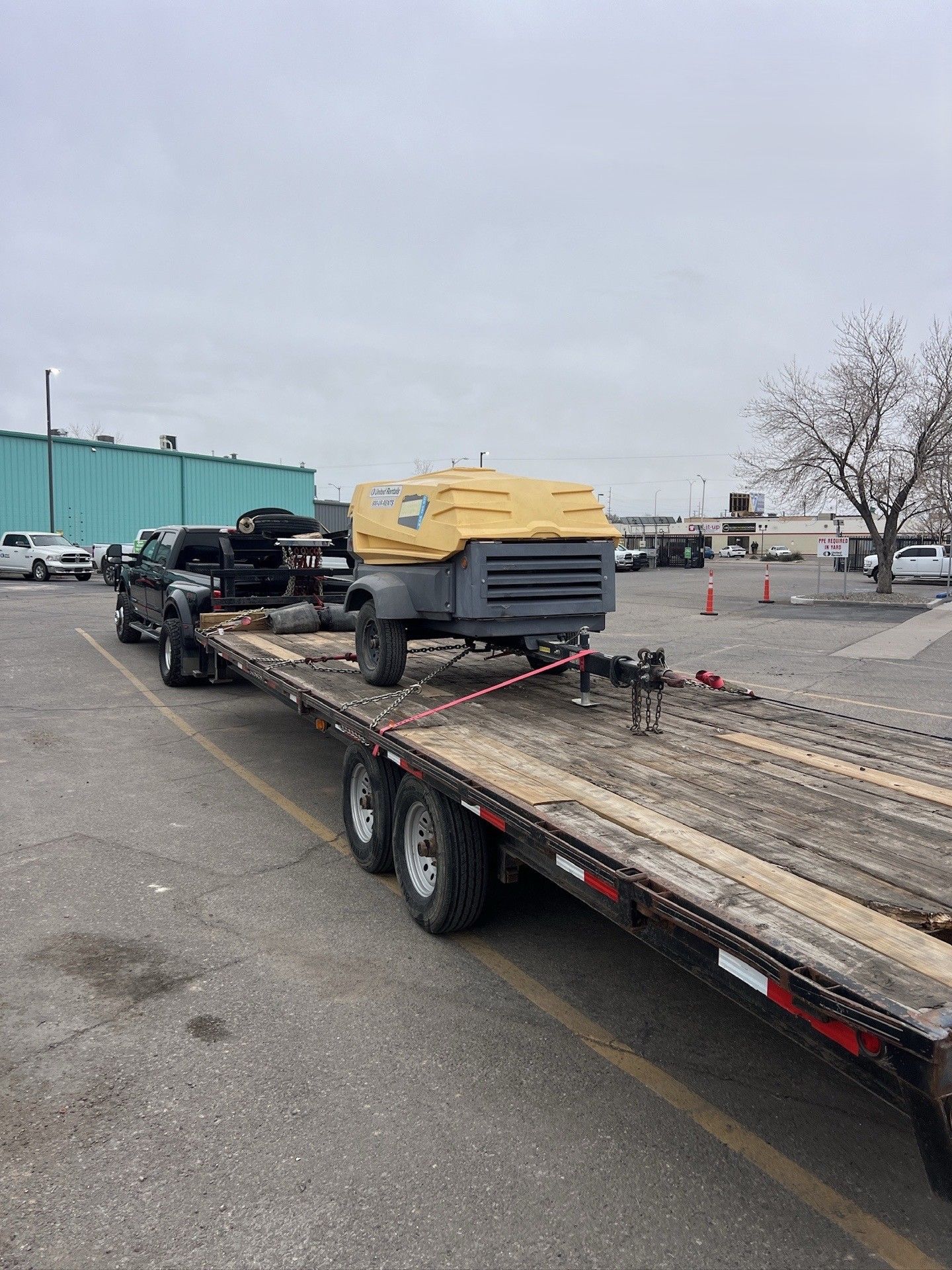 Yellow machine on a flatbed trailer in a parking lot with traffic cones and a teal building nearby