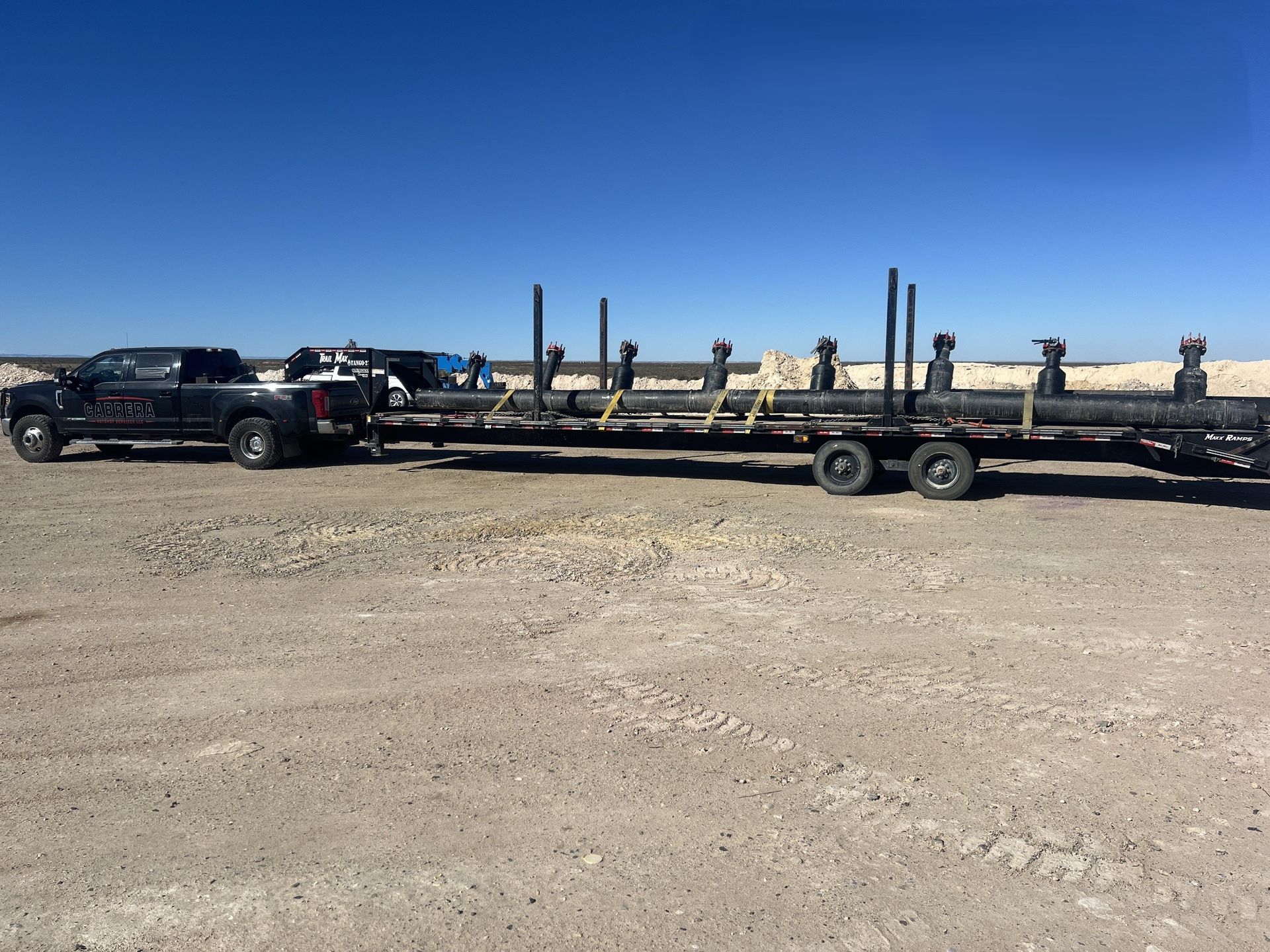 Black pickup truck towing a long flatbed trailer with equipment across a gravel lot under a blue sky