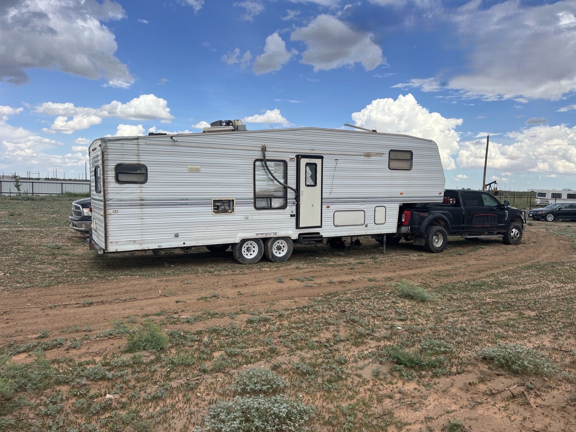White travel trailer hitched to a black pickup in a dirt lot under a cloudy blue sky.