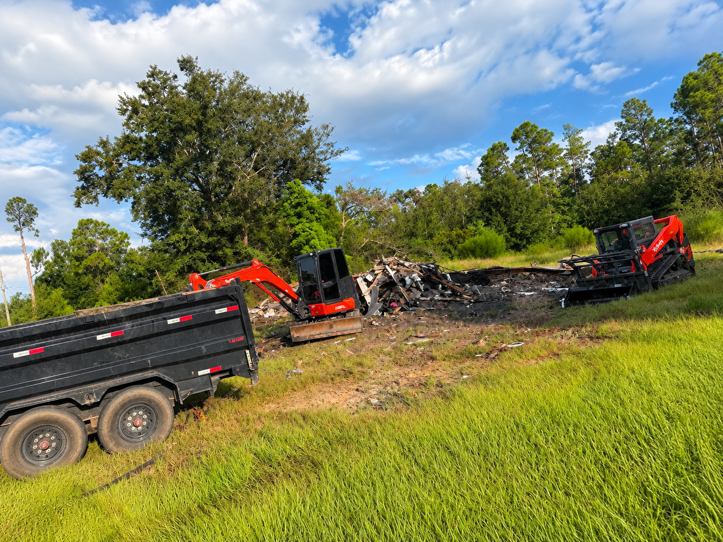 Orange excavators and a dump truck clearing debris in a grassy field under a cloudy blue sky