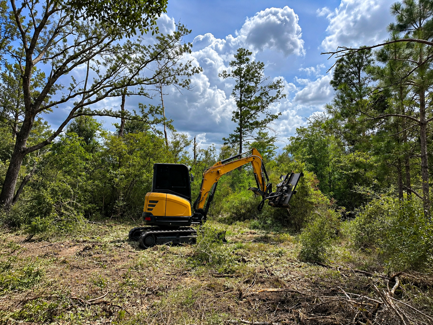Yellow excavator clearing brush in a wooded field under a cloudy sky